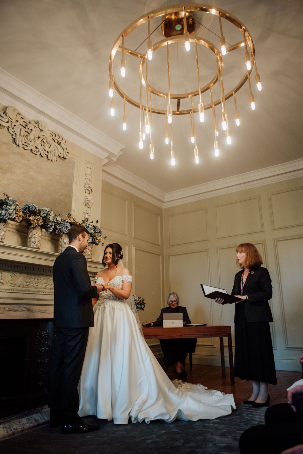 An elegant indoor wedding ceremony under a modern circular chandelier in a historic room.