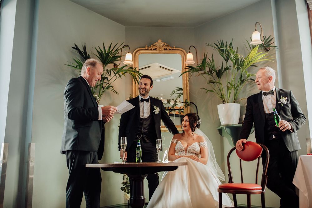 Guests celebrate with drinks and laughter in an elegant room with ornate mirror and tropical plants.