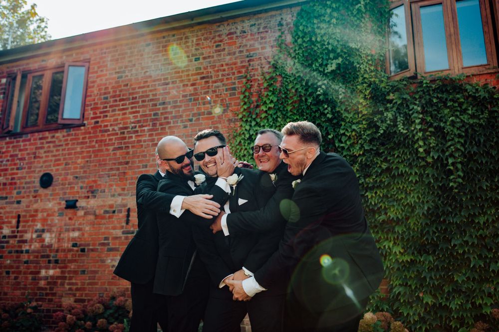 Groomsmen share a laugh together outside a rustic brick building covered in ivy.