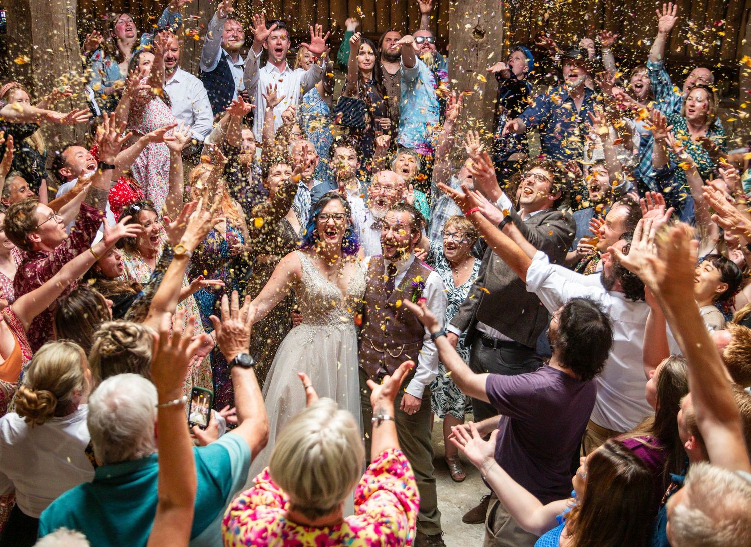 Large crowd of wedding guests dancing and celebrating together under string lights at an indoor venue.