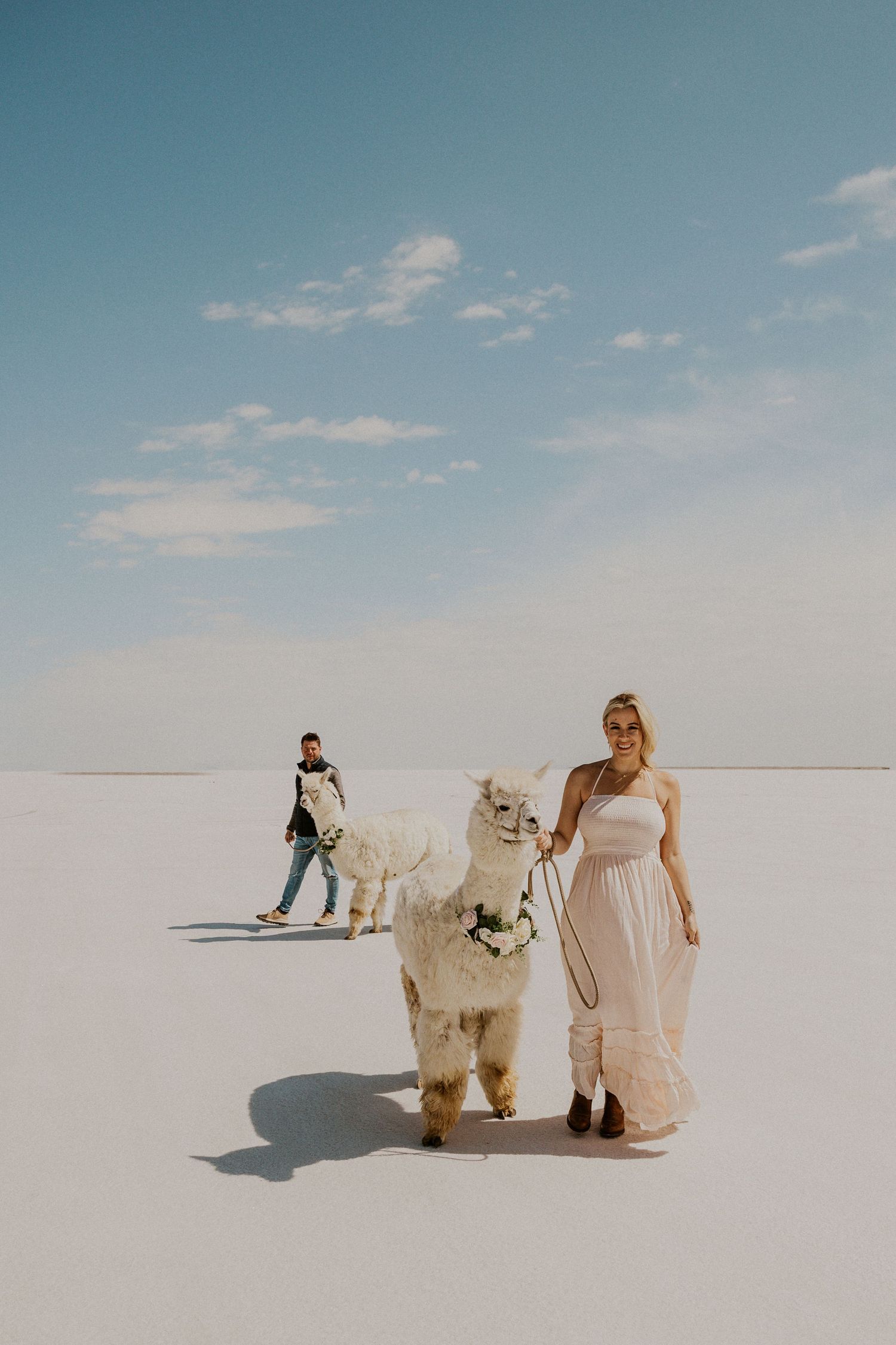 pregnant woman in a pink maxi dress walking an alpaca with floral necklace at Bonneville Salt Flats. Husband in background walking alpaca
