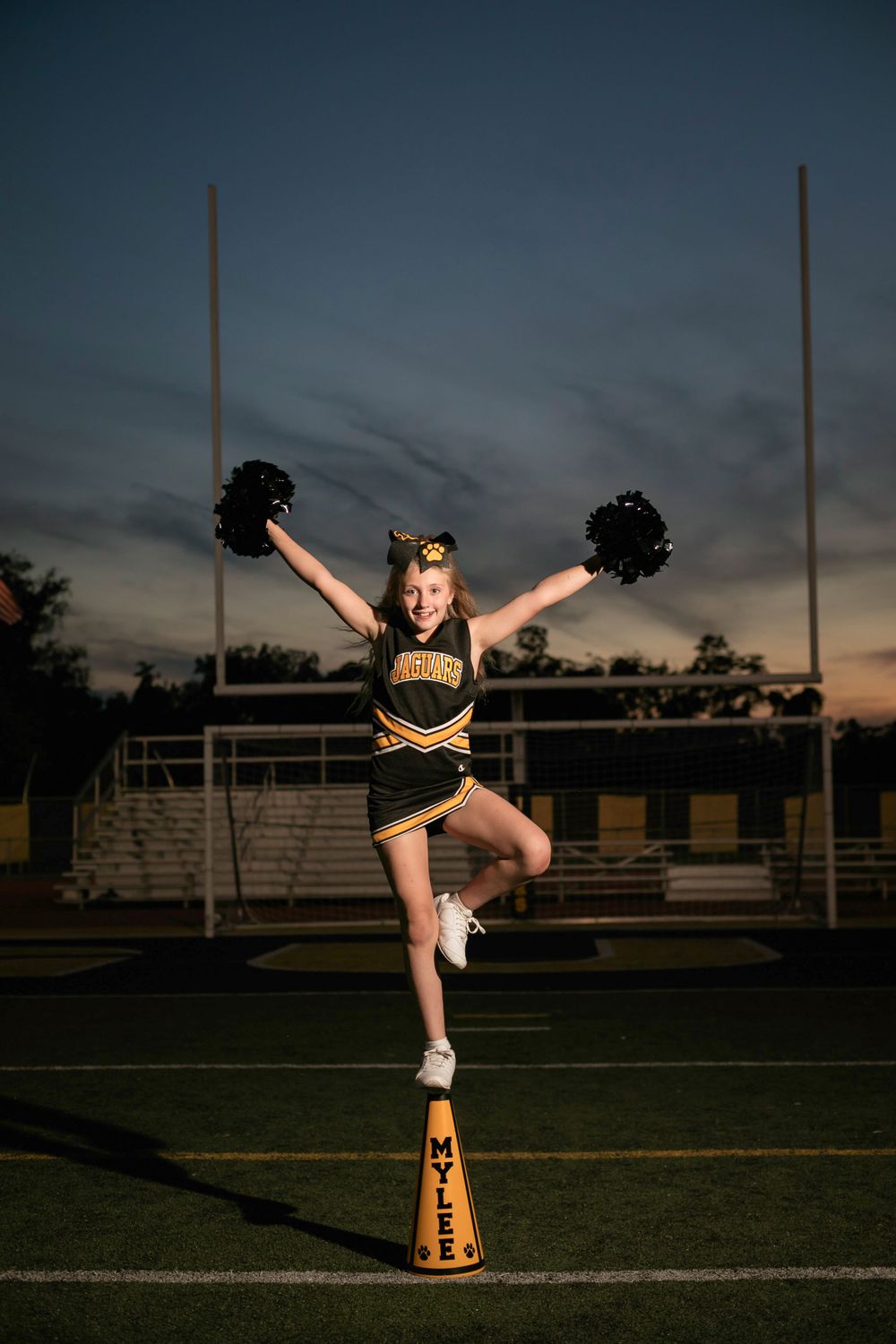 Cheerleader performs stunt on football field at dusk with raised pom poms.