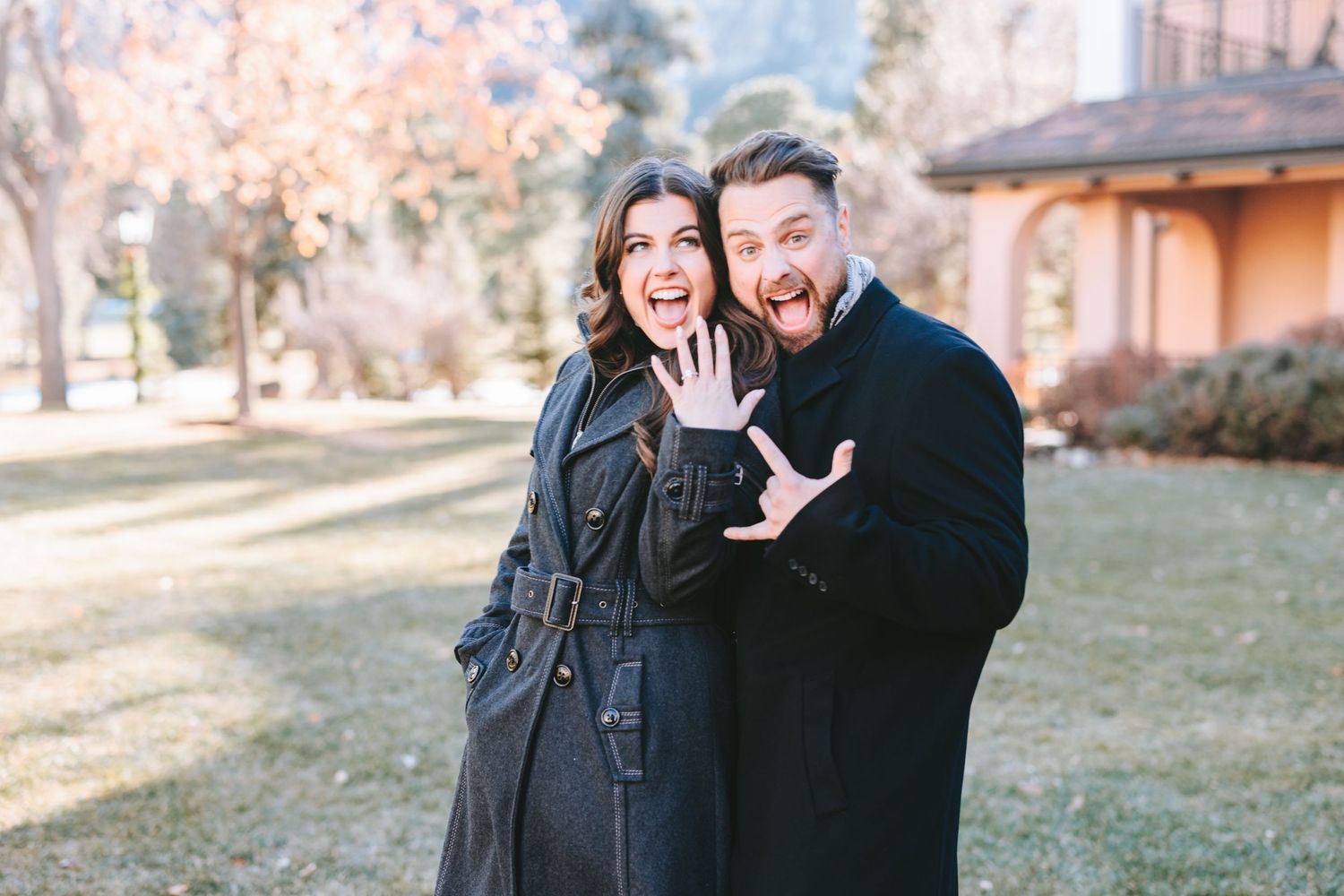 An excited couple showing off an engagement ring while wearing dark winter coats.
