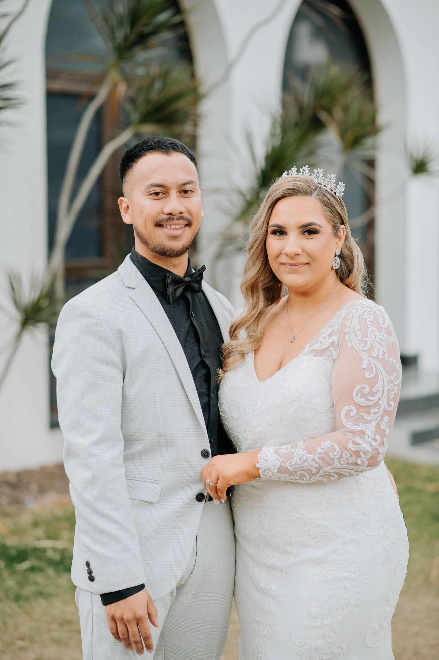 Wedding couple poses in front of white arched columns wearing elegant attire with bride in lace gown.
