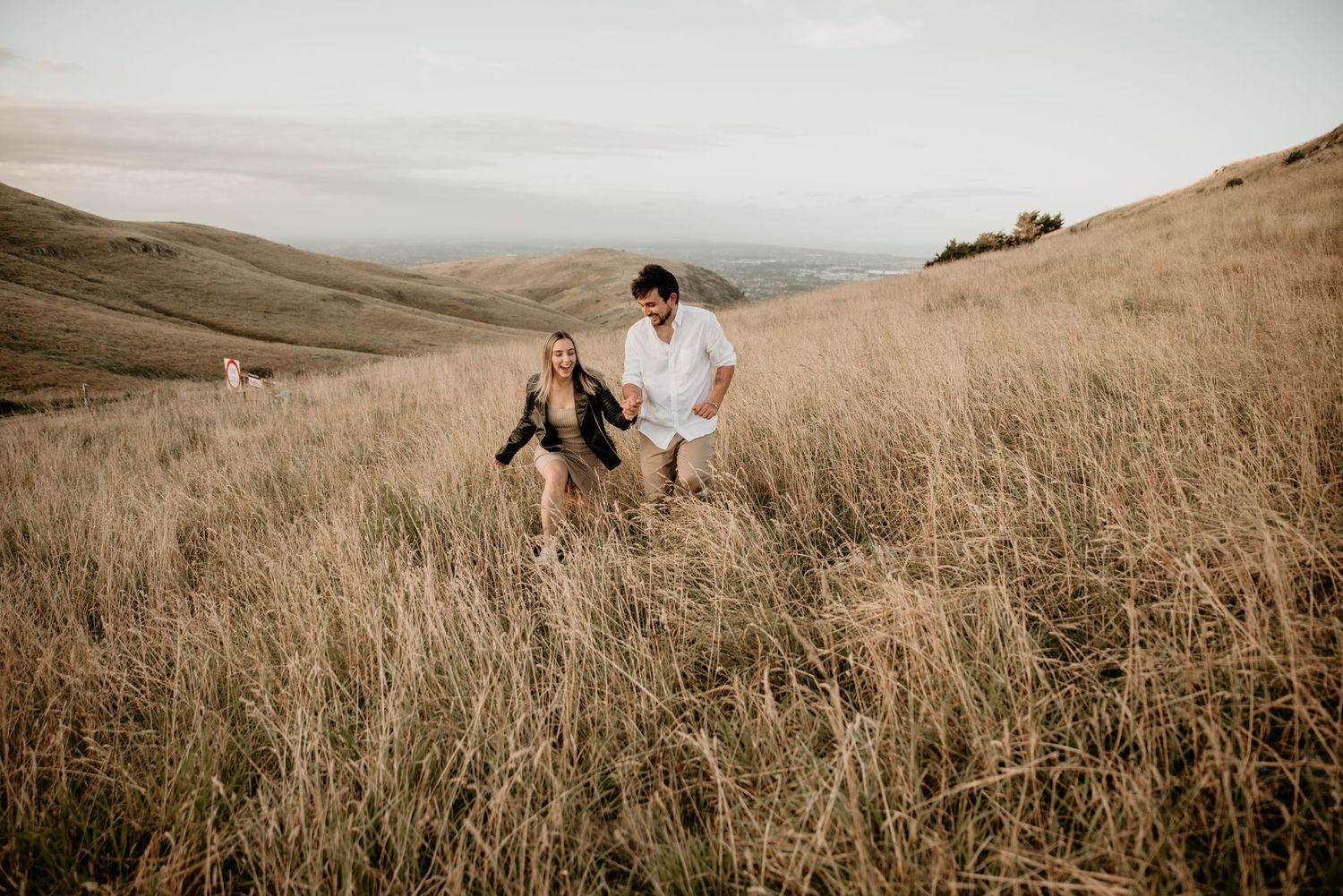 Couple walking through tall grass field during golden hour with mountain views.