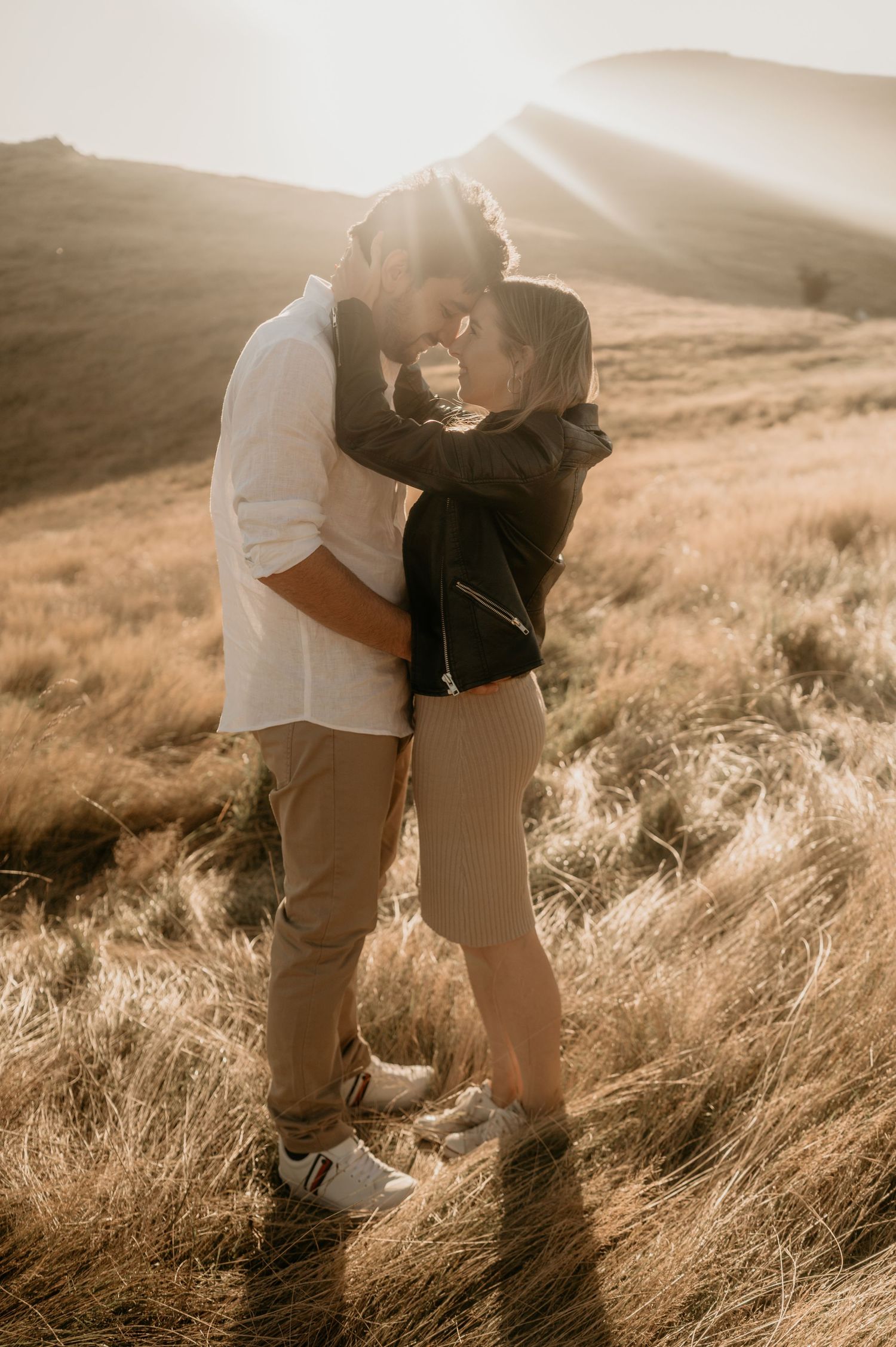 Intimate moment shared between couple in tall grass with sunlit mountains behind them.
