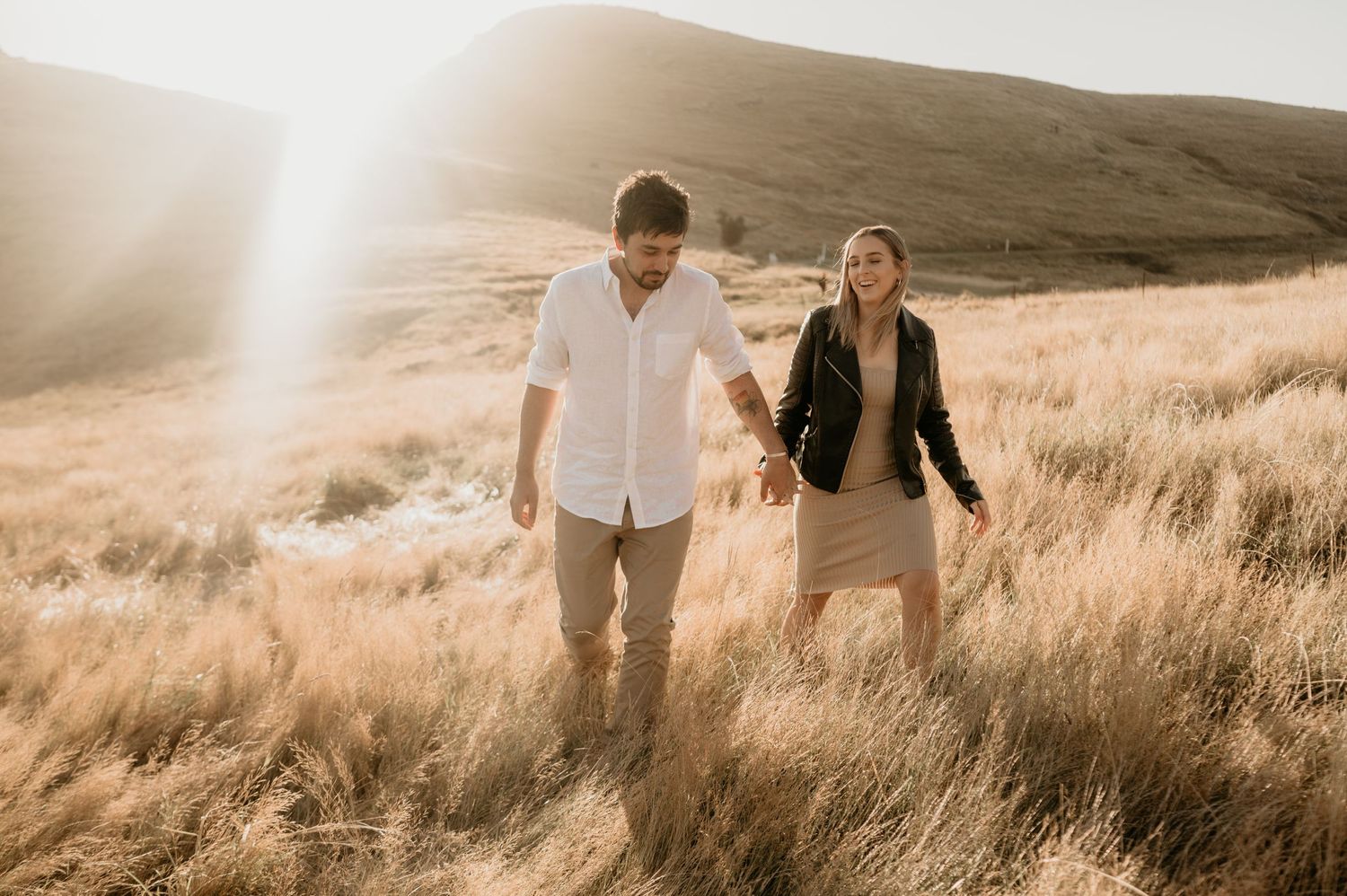 Couple walking hand in hand through sunlit mountain meadow at golden hour.