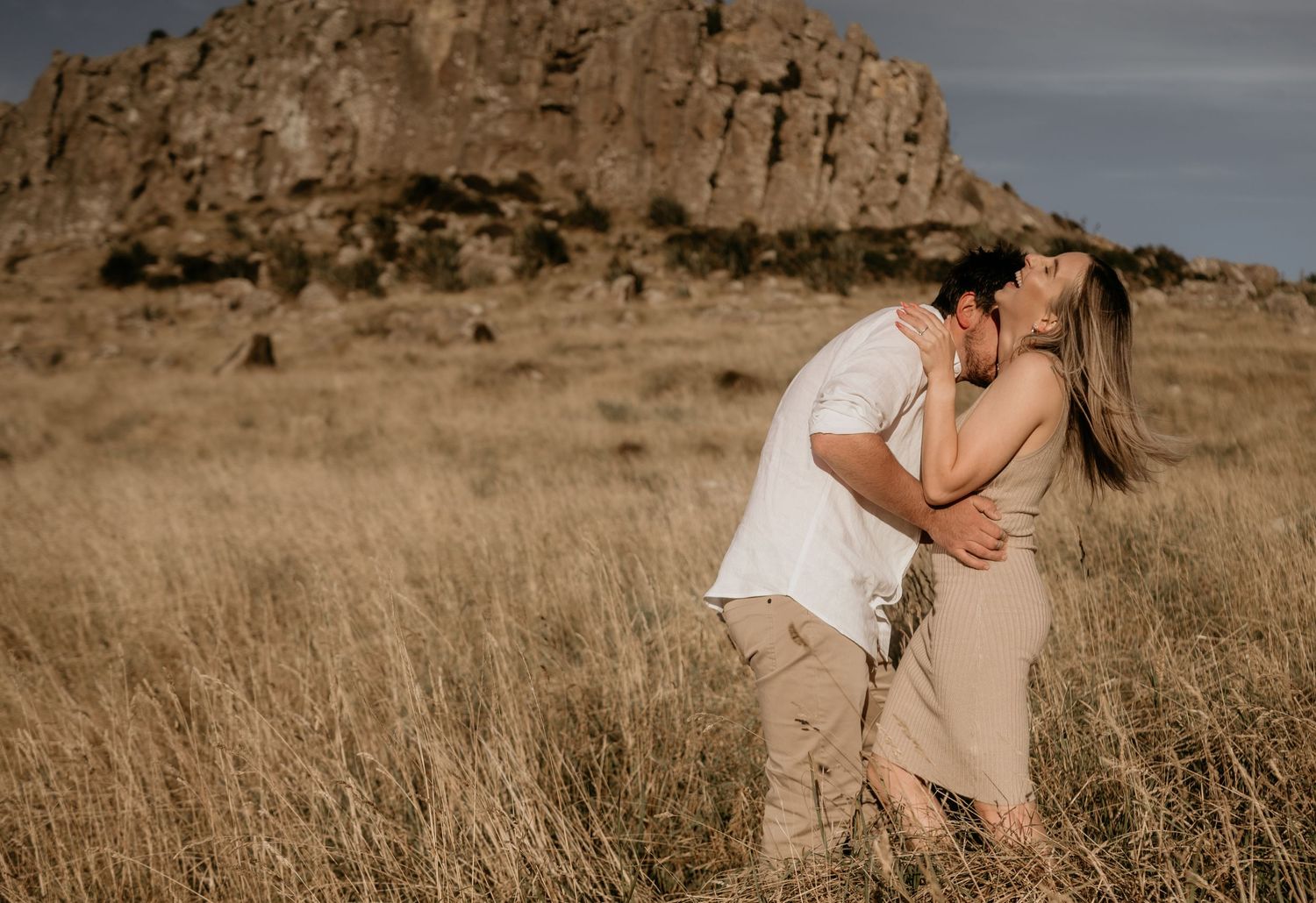 Romantic couple sharing intimate moment in dried grass field with rocky mountain backdrop.