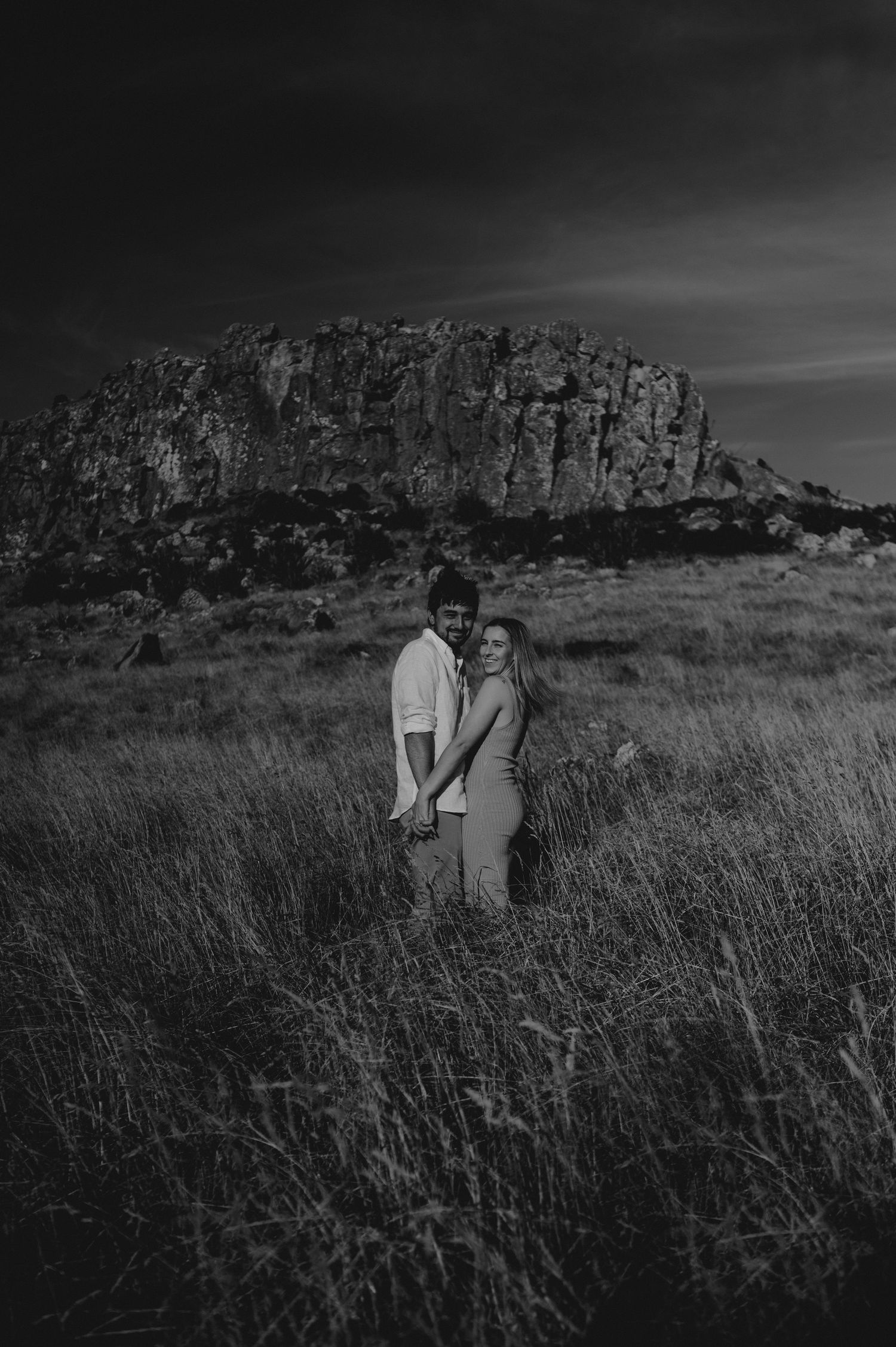 Black and white photo of couple standing in tall grass with dramatic rocky cliff behind them.