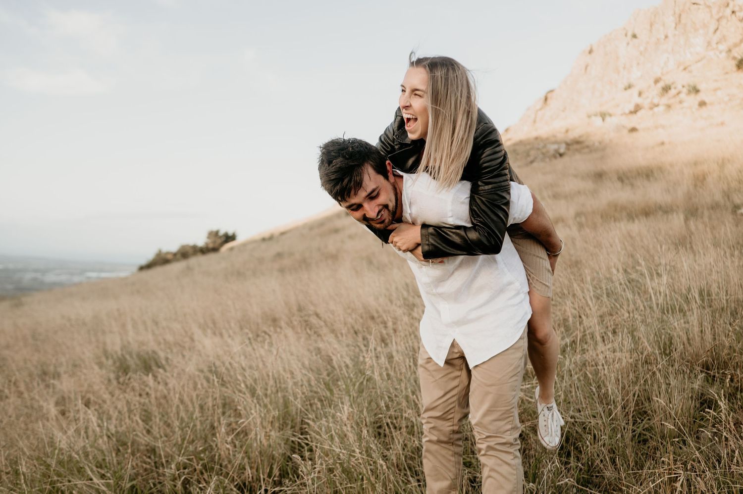 Playful couple enjoying a piggyback ride in a golden hillside field during sunset.