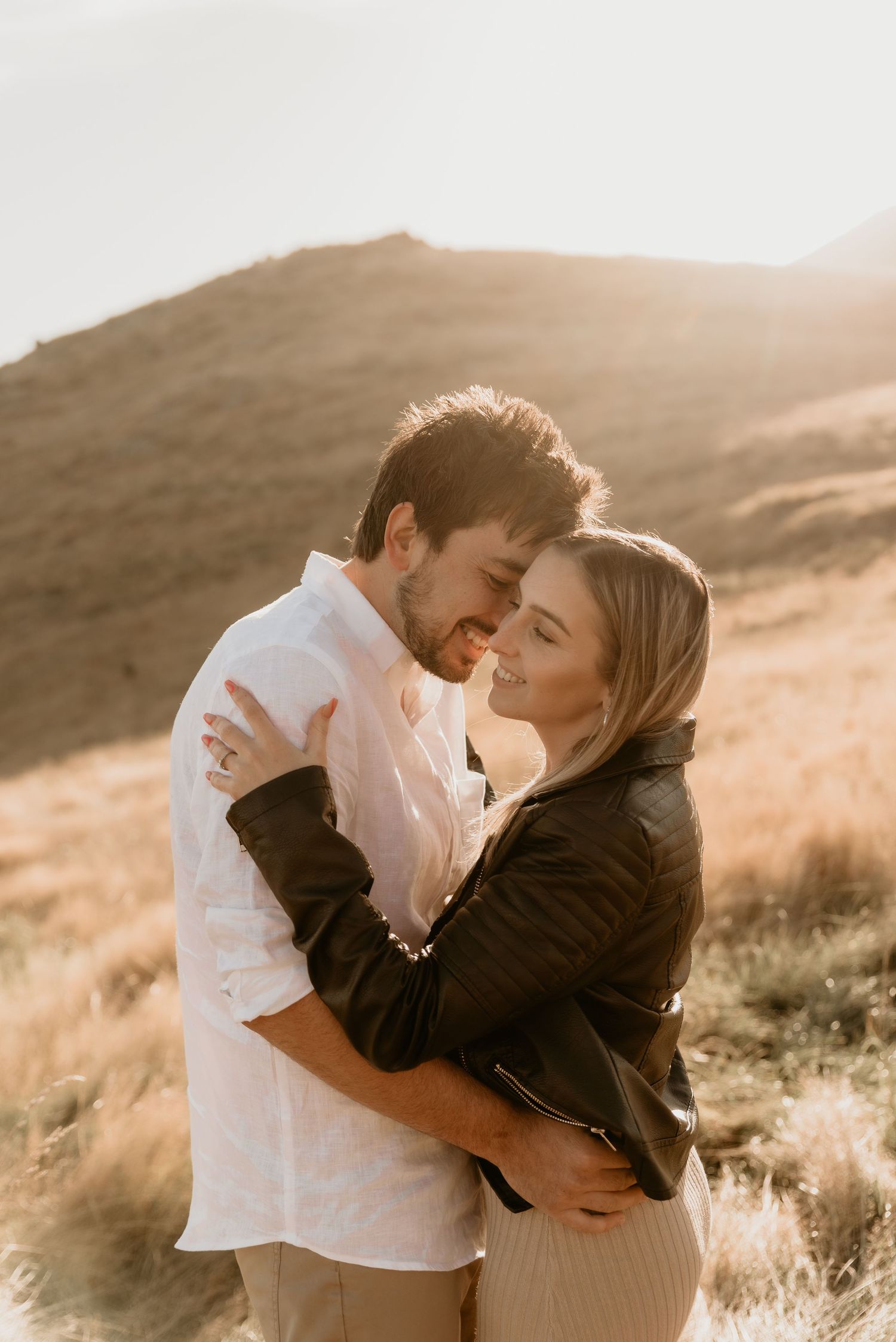 Romantic sunset engagement photo with couple embracing against mountain landscape.
