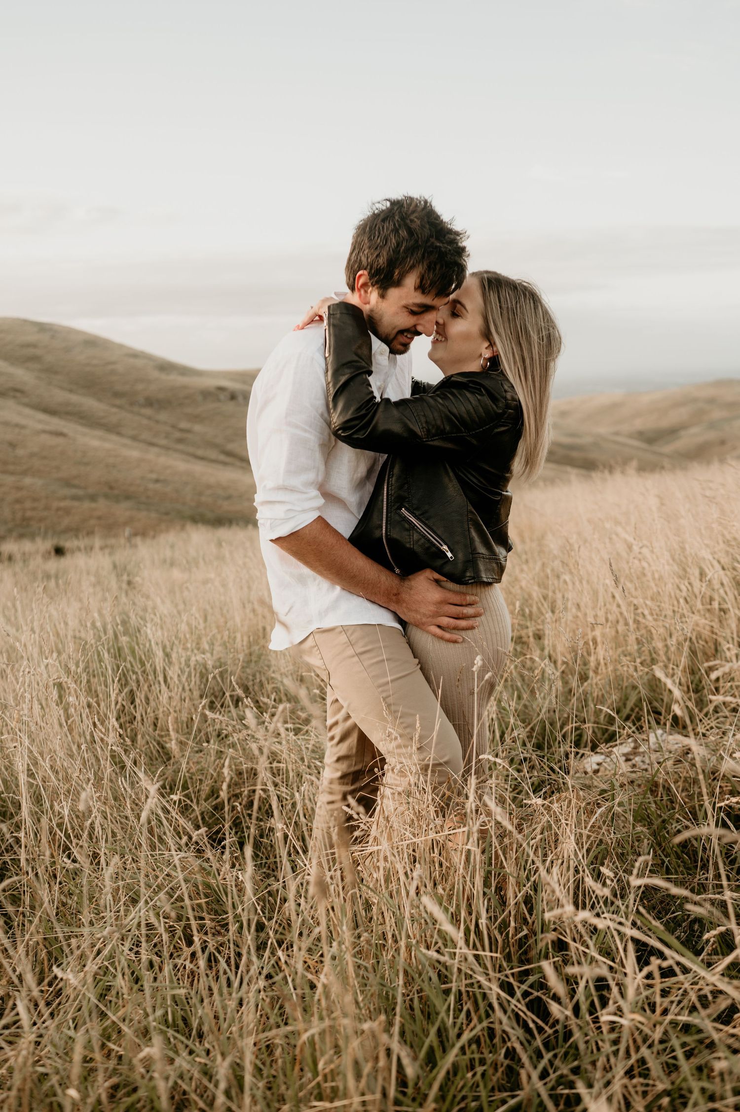 Romantic embrace in tall grass with soft sunlight and rolling hills in background.