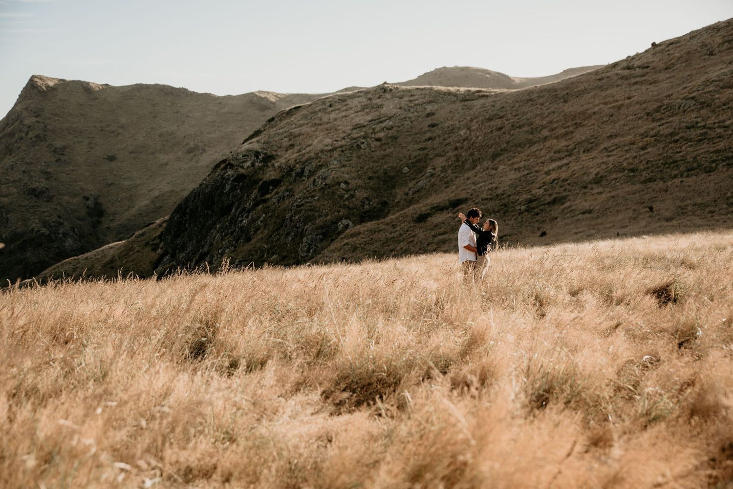 Couple walking together on a scenic mountain trail through golden grass fields at sunset.