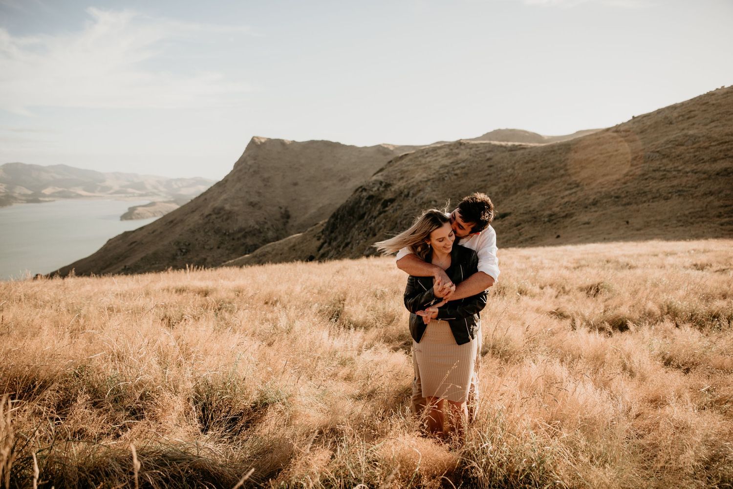 Couple embracing in golden meadow with dramatic coastal mountain views.