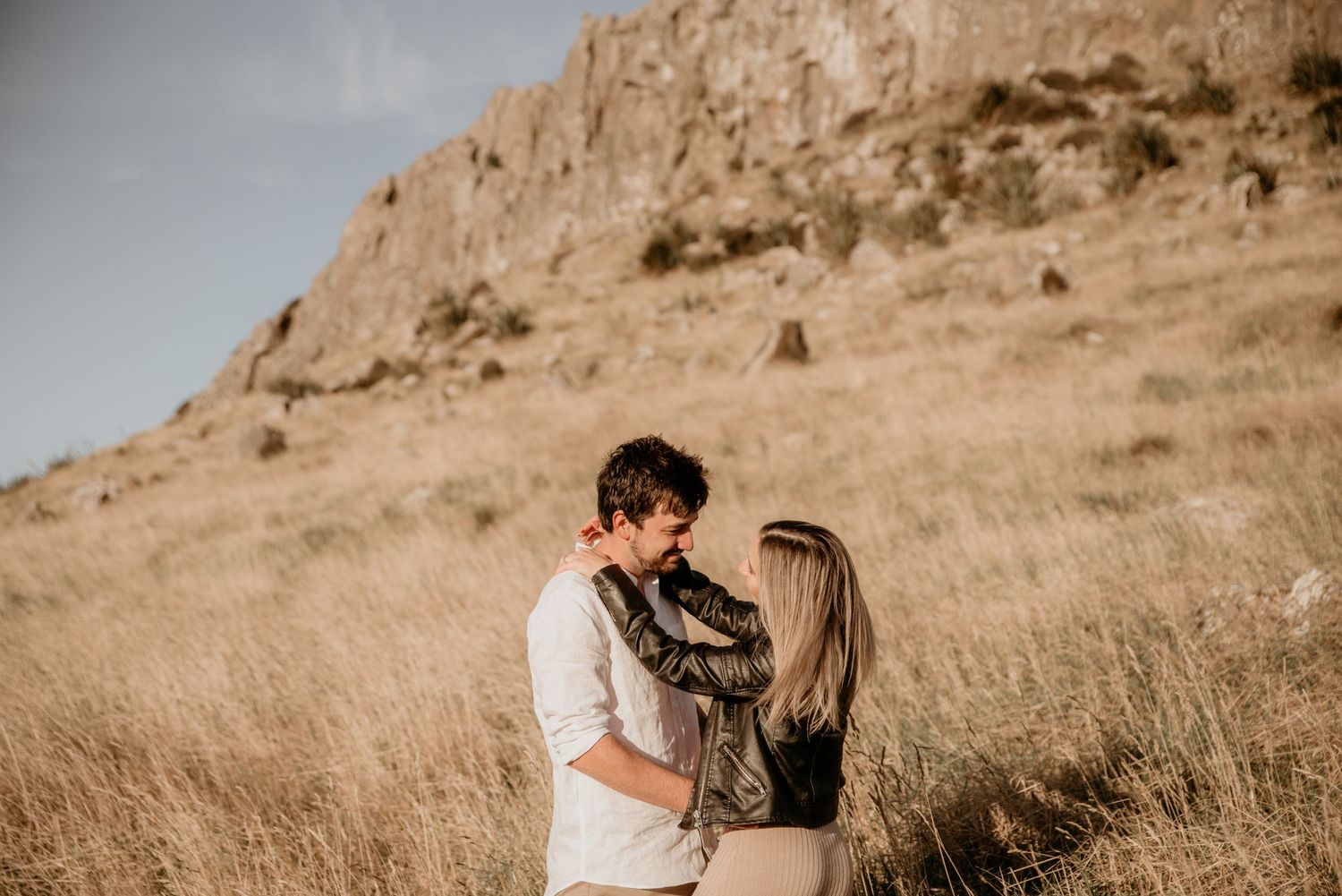 Loving couple shares tender moment against backdrop of rugged cliffs and dry grass.