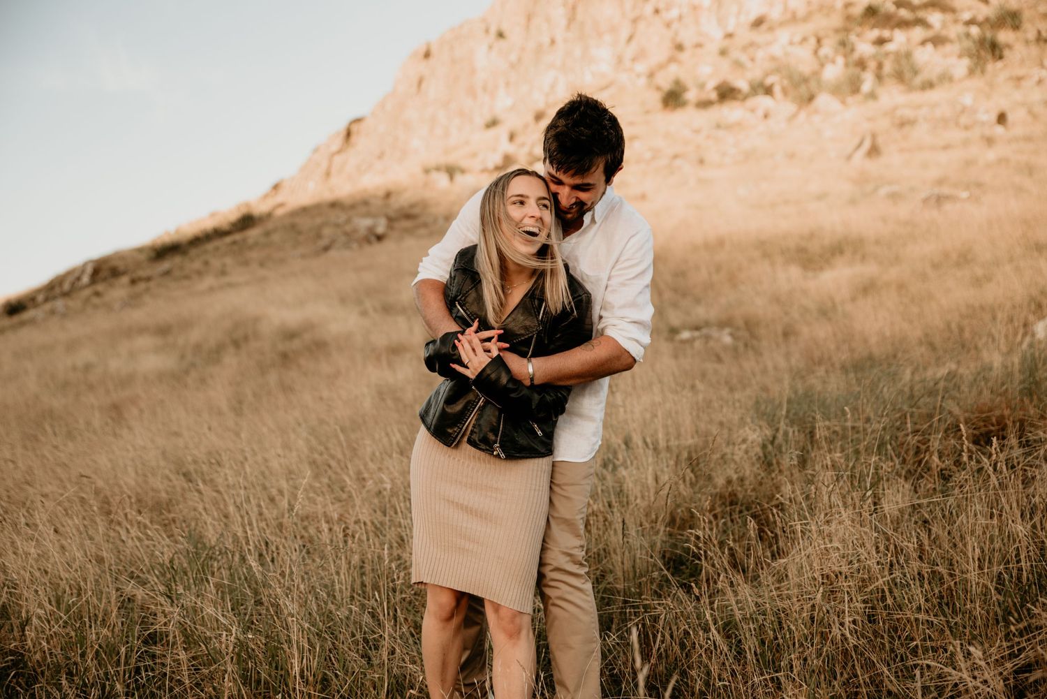 Sweet couple embracing while standing in a golden mountain meadow at sunset.