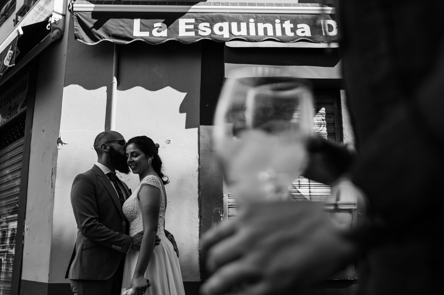 Black and white photo of a couple embracing outside La Esquinita storefront while a blurred figure watches in Seville, Spain