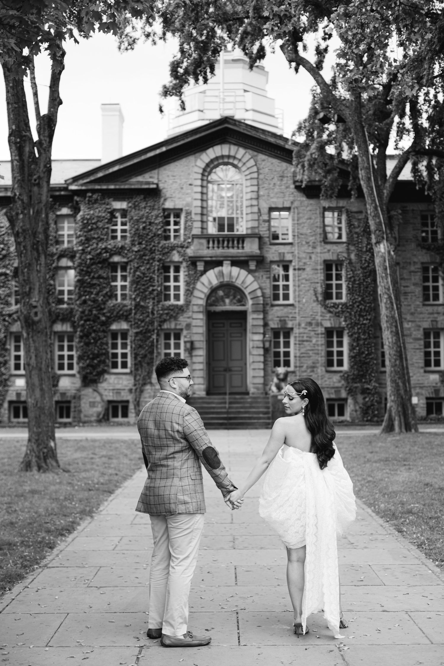 A couple holding hands in front of a historic stone building with trees on a university campus.