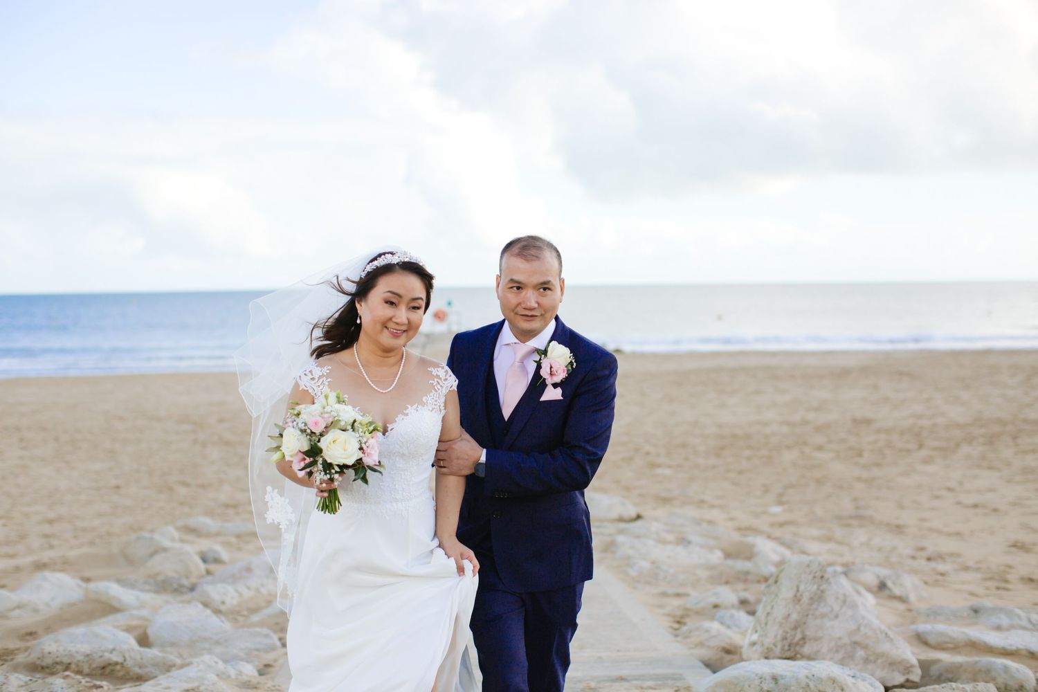 A couple in wedding attire pose on a beach with rocks and ocean in the background.