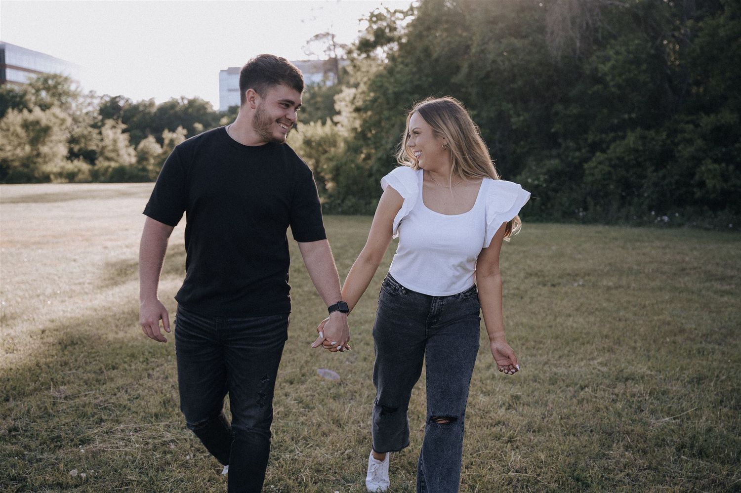 Couple in casual attire walking hand in hand through a sunlit field.