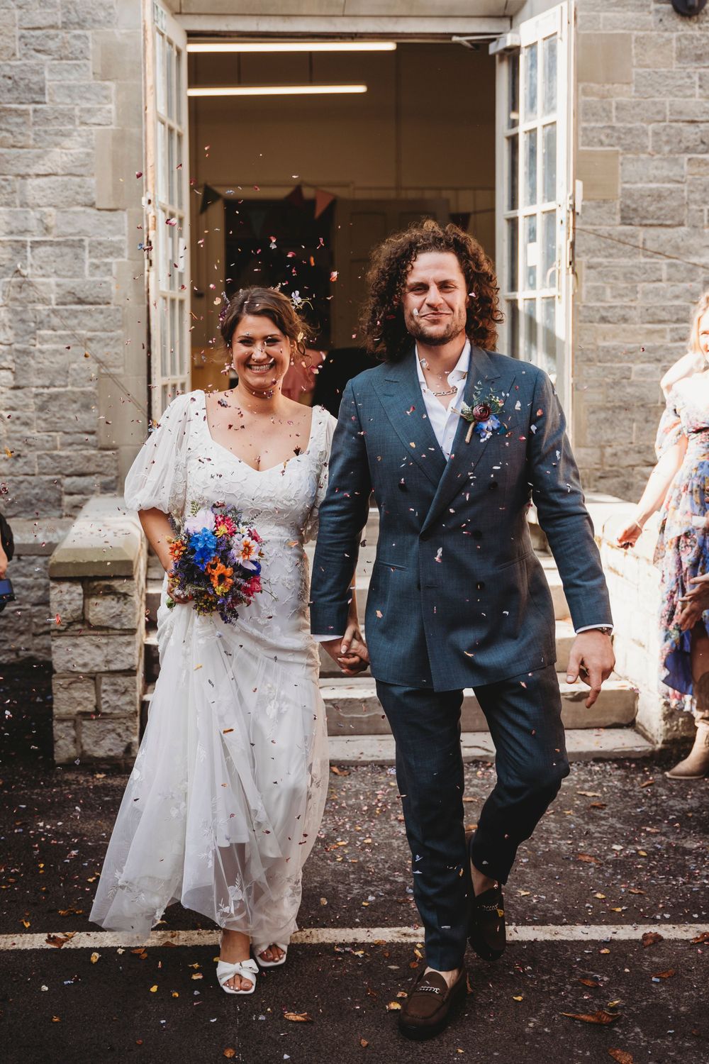 Newlyweds walking through confetti shower outside historic stone building entrance.