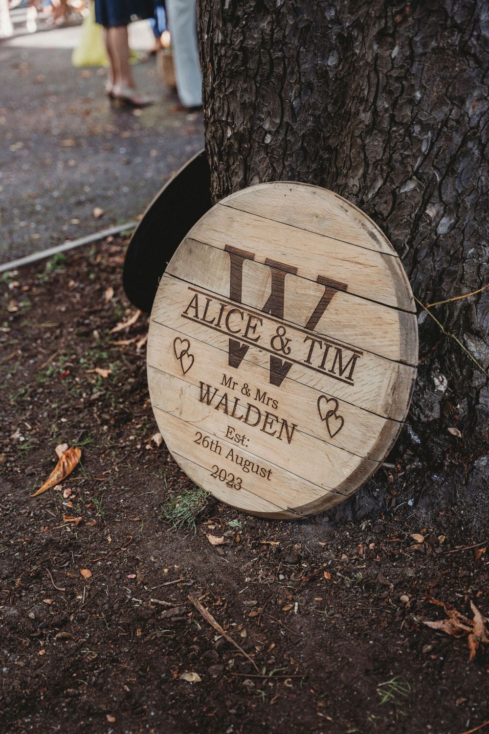 Wooden circular wedding sign with names and date engraved leaning against a tree trunk.