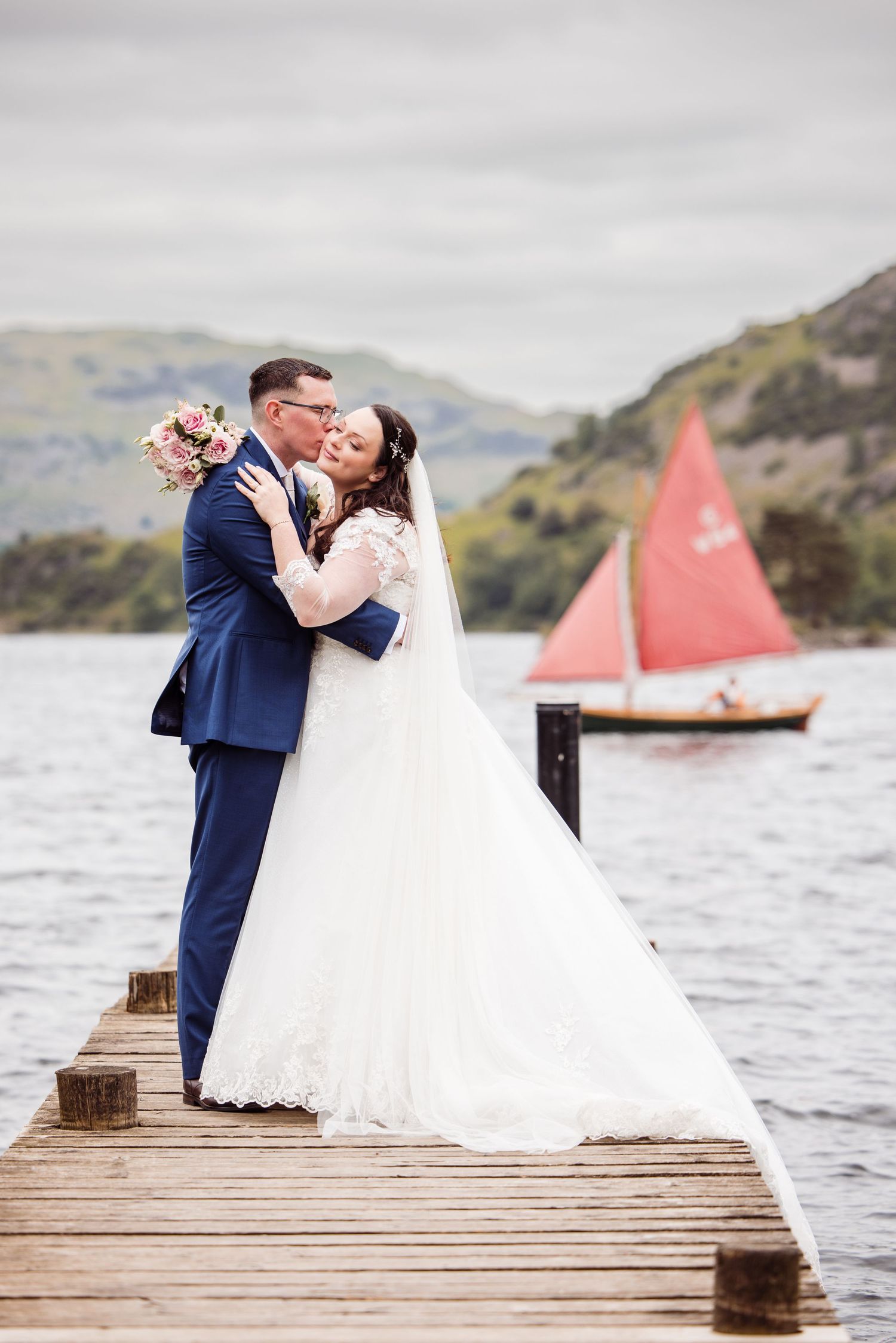 A wedding couple embraces on a lakeside dock with a red-sailed boat in the background.