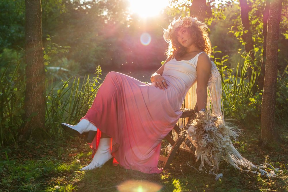 April Pearson sat leaning near a tree wearing pink boho wedding dress with a floral bouquet during sunset golden hour.