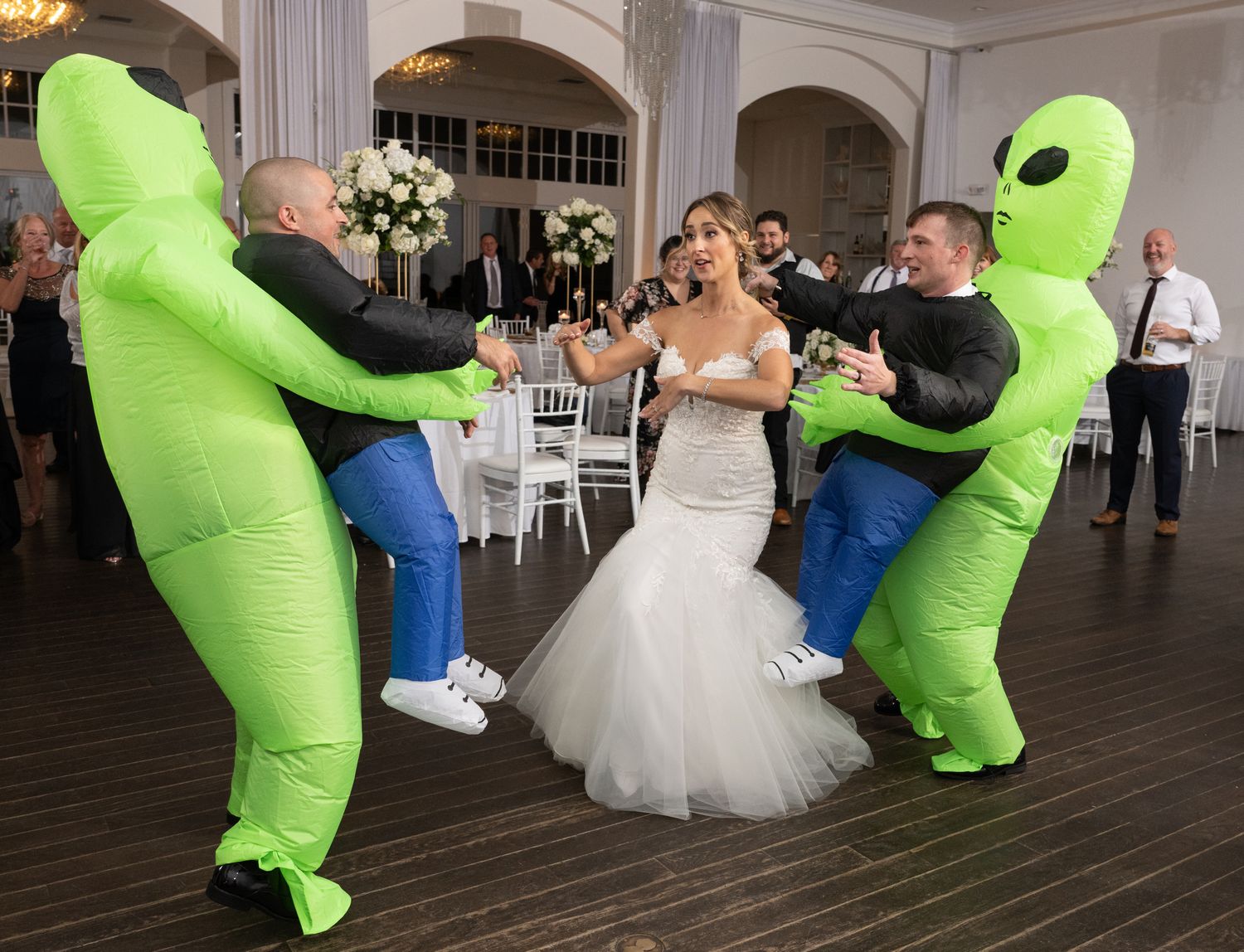 People in inflatable green alien costumes dancing with a bride at a wedding reception.