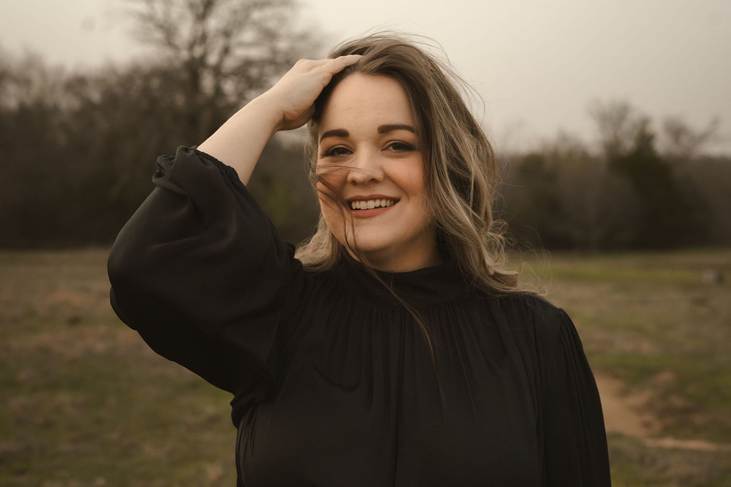 a woman in an open landscape wearing a black dress smiling at you while she pushes hair out of her face