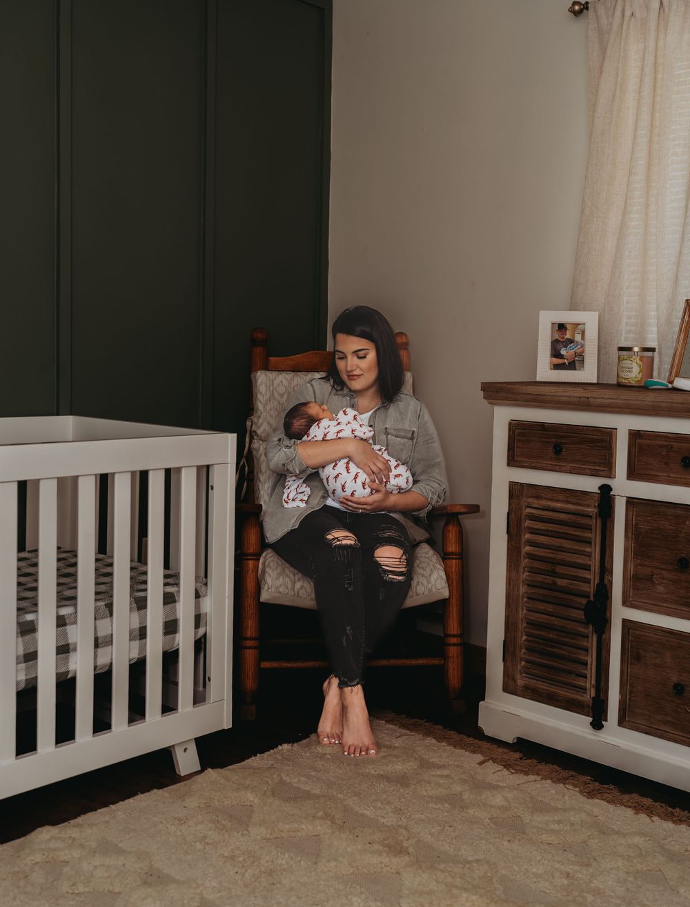 Mother in rocking chair holding sleeping newborn in a nursery with white crib.