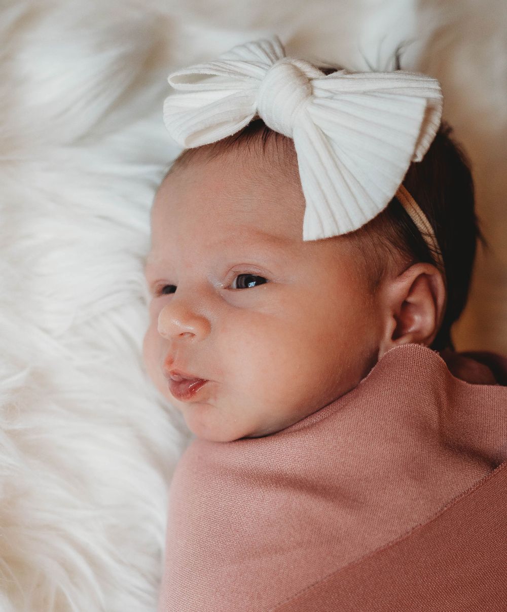 Baby wearing large white bow headband wrapped in pink blanket on white bedding.