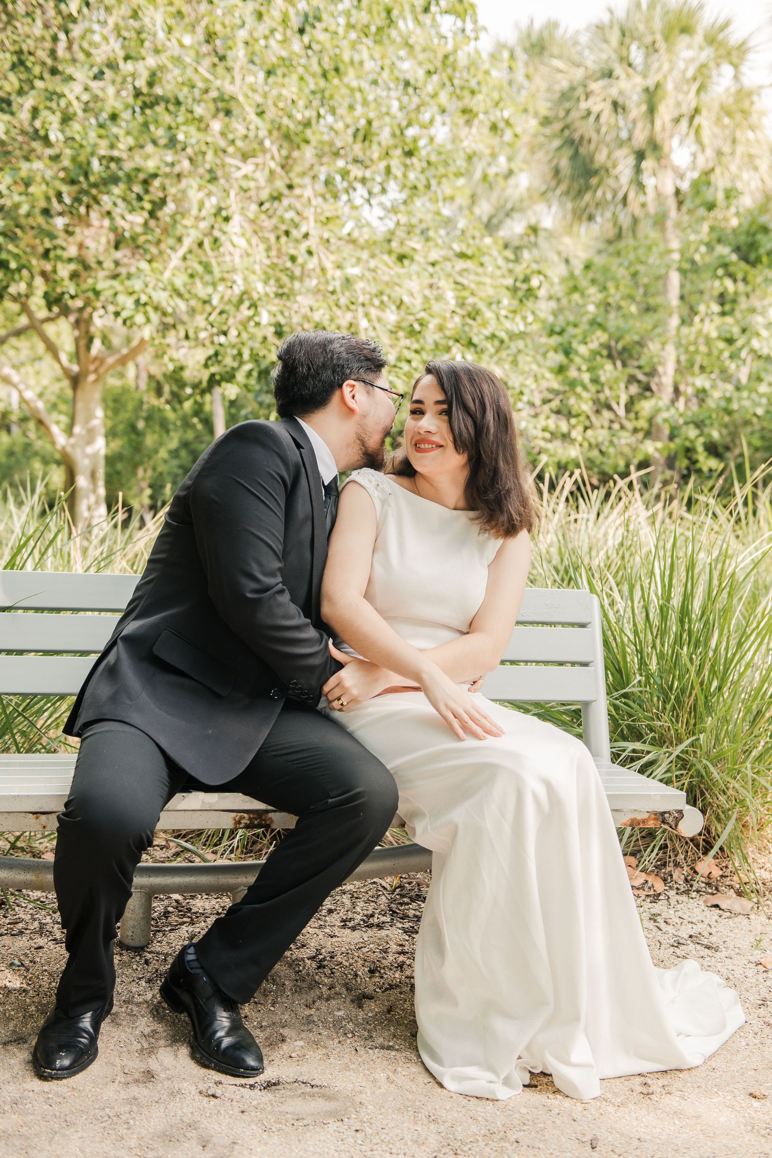 Newlywed couple embracing on wooden bench in garden setting with palm trees in background.