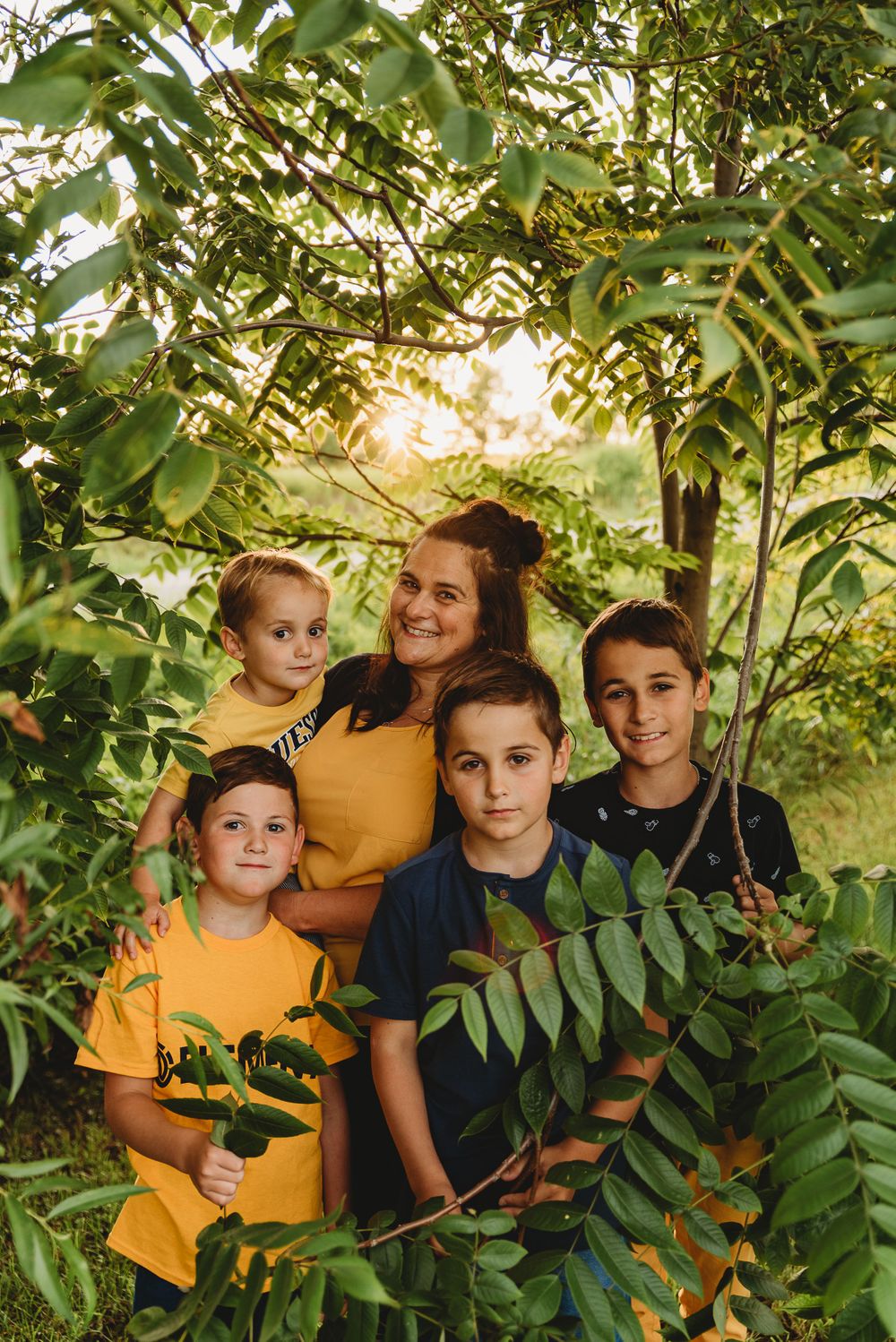 Family group poses among leafy green trees in natural outdoor setting at sunset.