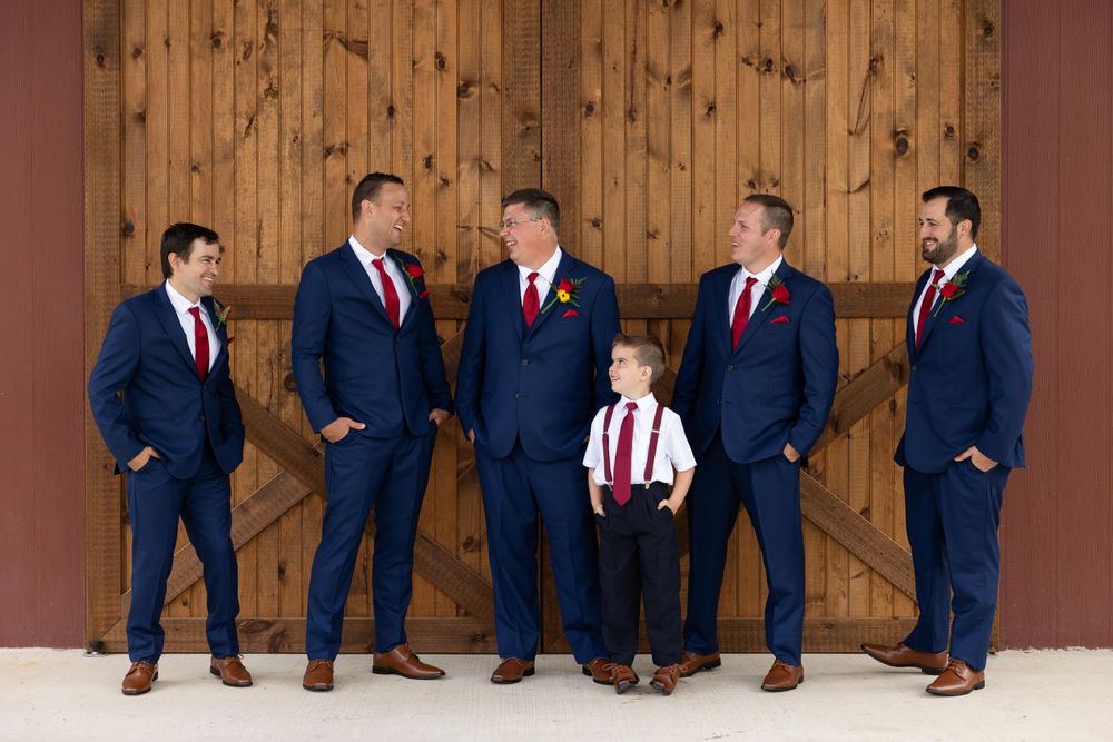 A wedding party in navy suits with red ties poses against a wooden barn door background.