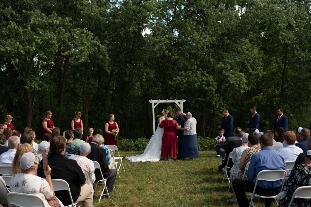 An outdoor wedding ceremony under a white arch with guests seated on folding chairs amid green trees.