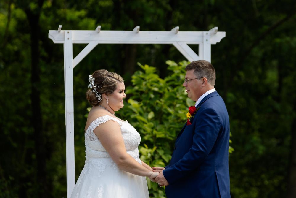 A couple exchanges vows under a white pergola during an outdoor wedding ceremony.