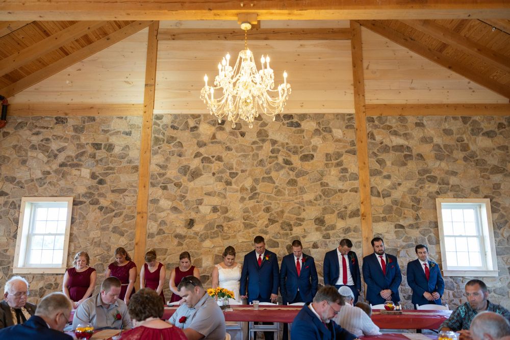 Wedding party and guests gathered in a rustic stone-walled venue with wooden beams and crystal chandelier.