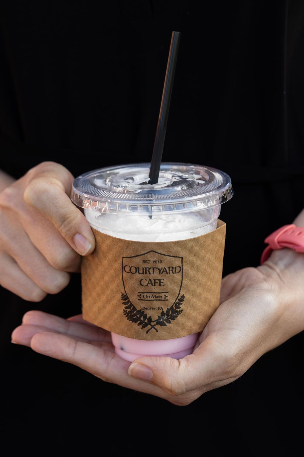 Hands holding a drink from Courtyard Cafe in a clear plastic cup with brown sleeve.