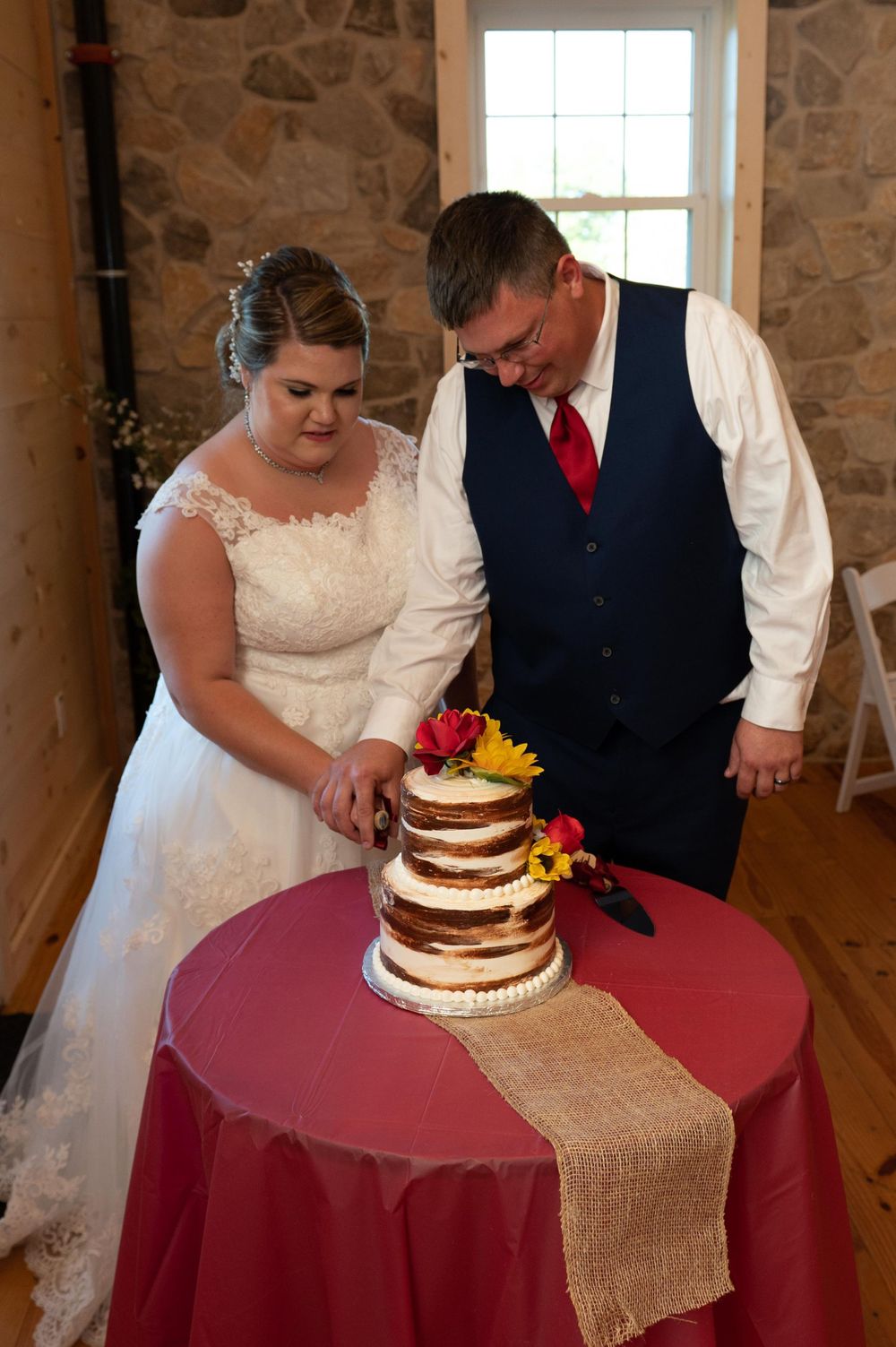 A couple cutting their rustic naked wedding cake decorated with yellow and red flowers on a burgundy tablecloth.