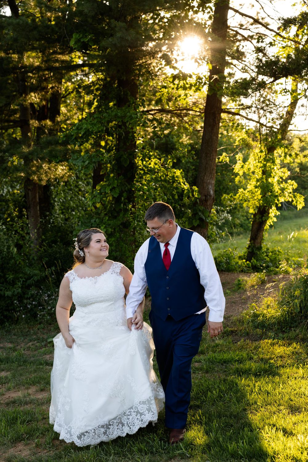 Wedding couple walking hand in hand through a sunlit wooded area in formal attire.