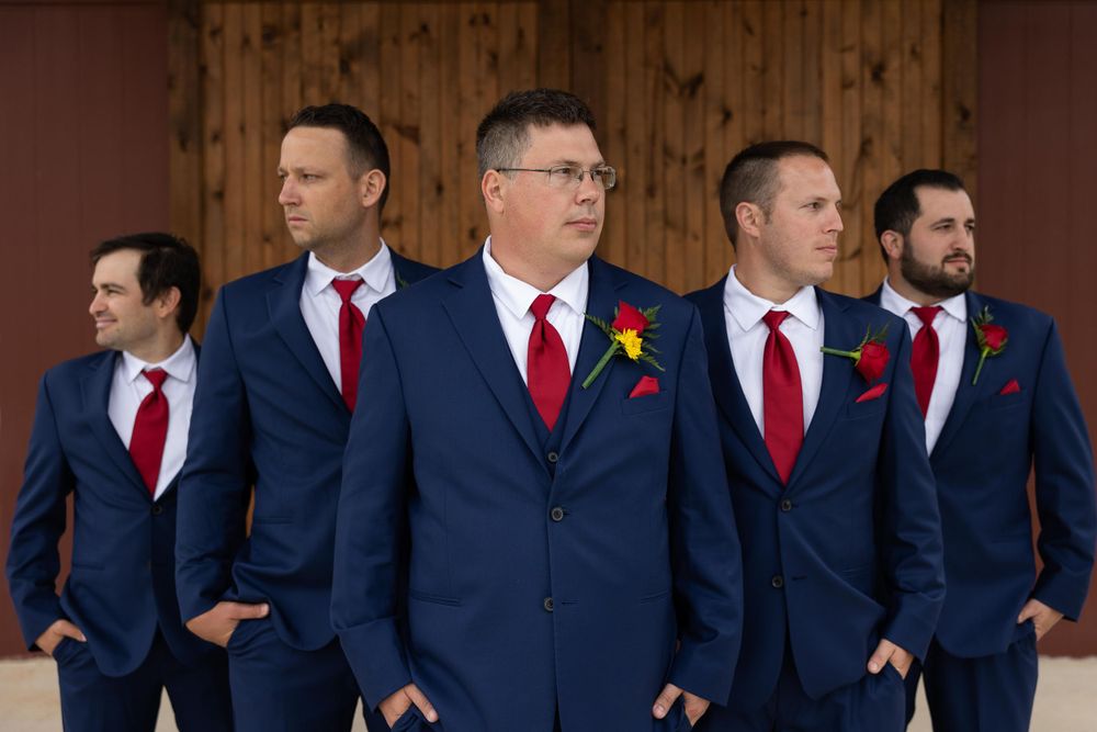 Groomsmen in matching navy suits and red ties stand in a line against wooden paneling.