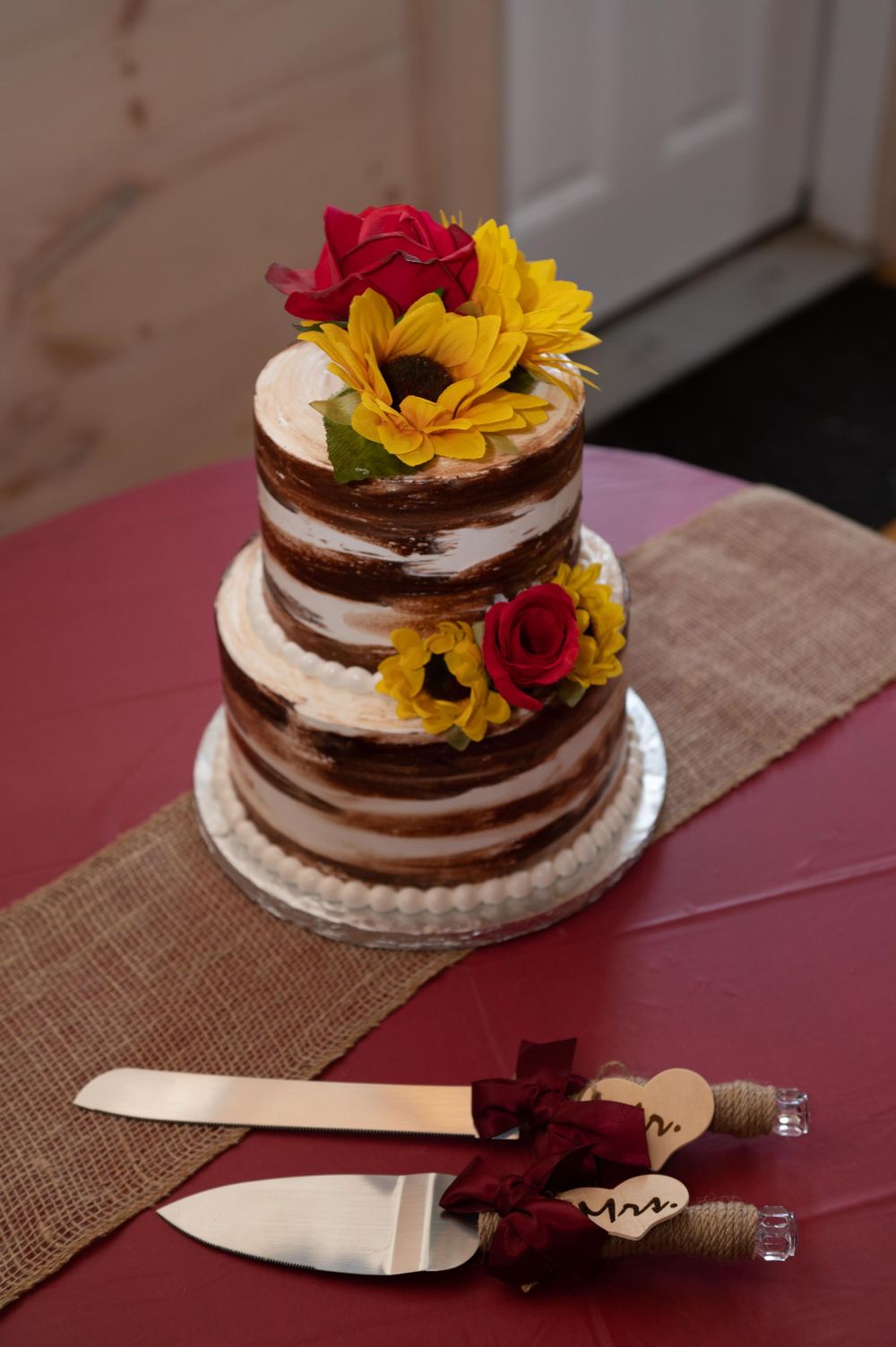A rustic naked wedding cake decorated with red roses and yellow sunflowers on a burlap runner.