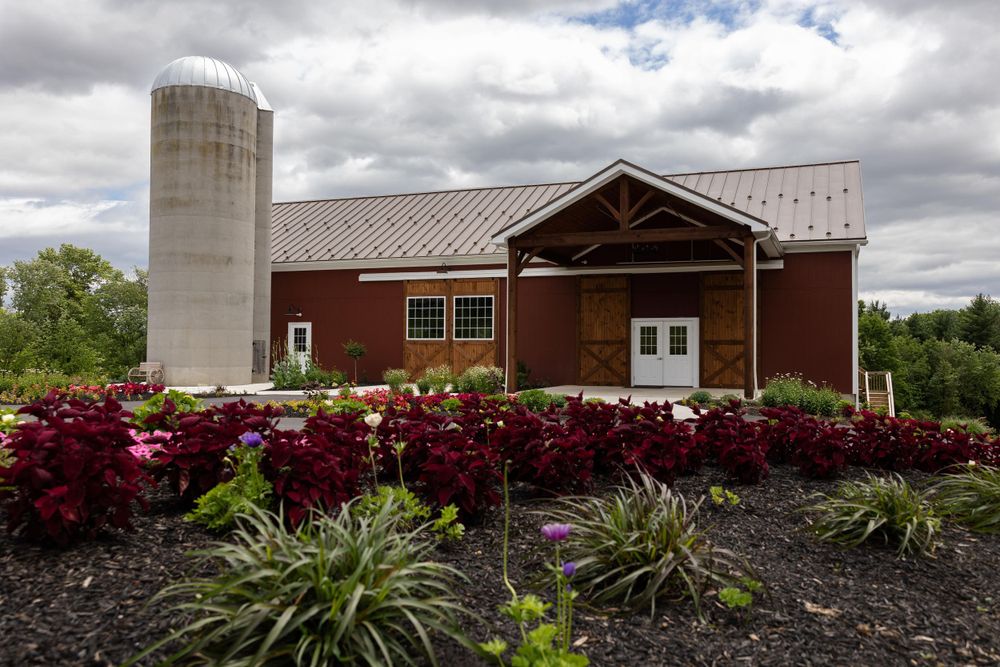A red barn with silo and landscaped garden featuring burgundy flowers in the foreground.