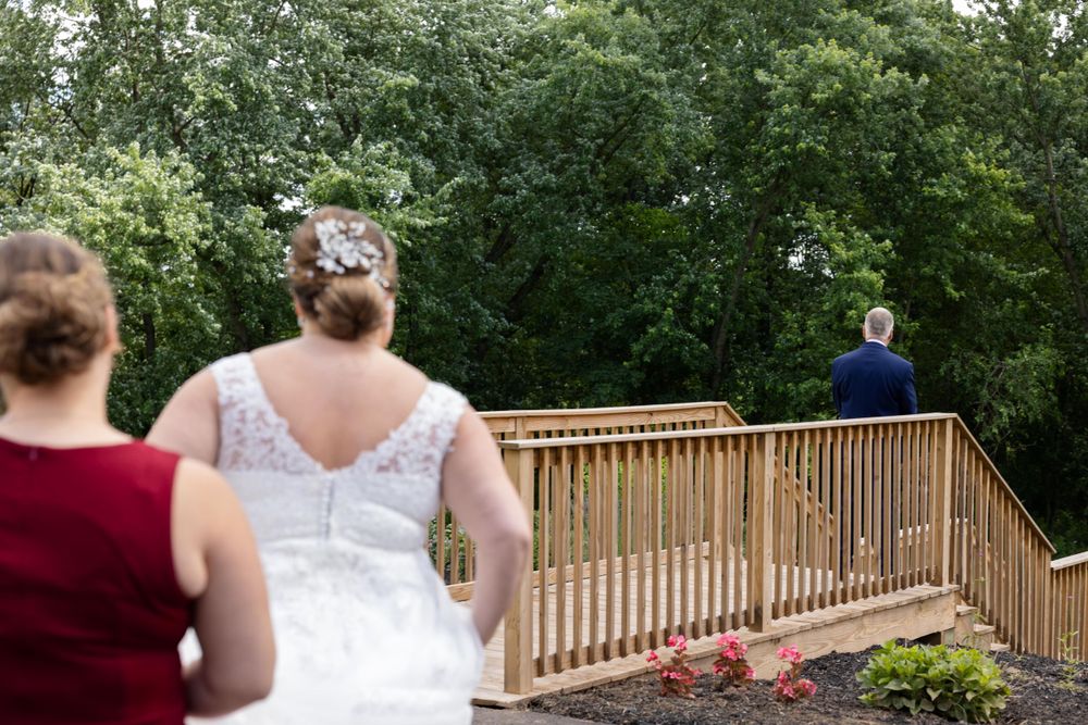 Wedding photography captures a first look moment on a wooden deck surrounded by lush green trees.