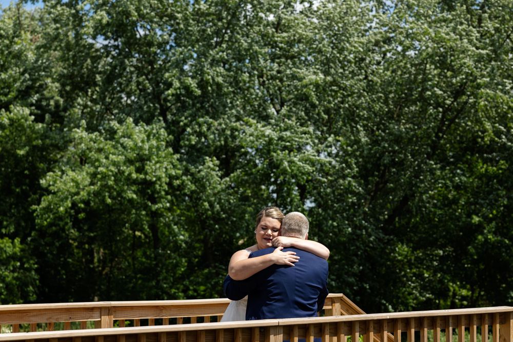 A heartfelt embrace is shared on a wooden deck with sunlight streaming through the trees.