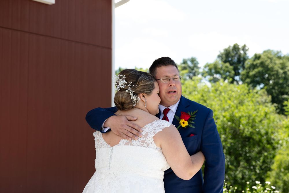 An emotional wedding moment is captured in front of a rustic red barn wall.