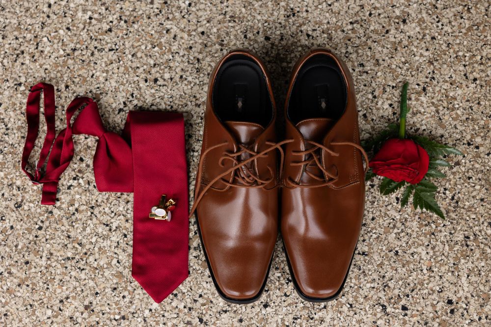 Wedding accessories including brown dress shoes, red tie, and rose laid out on carpet.