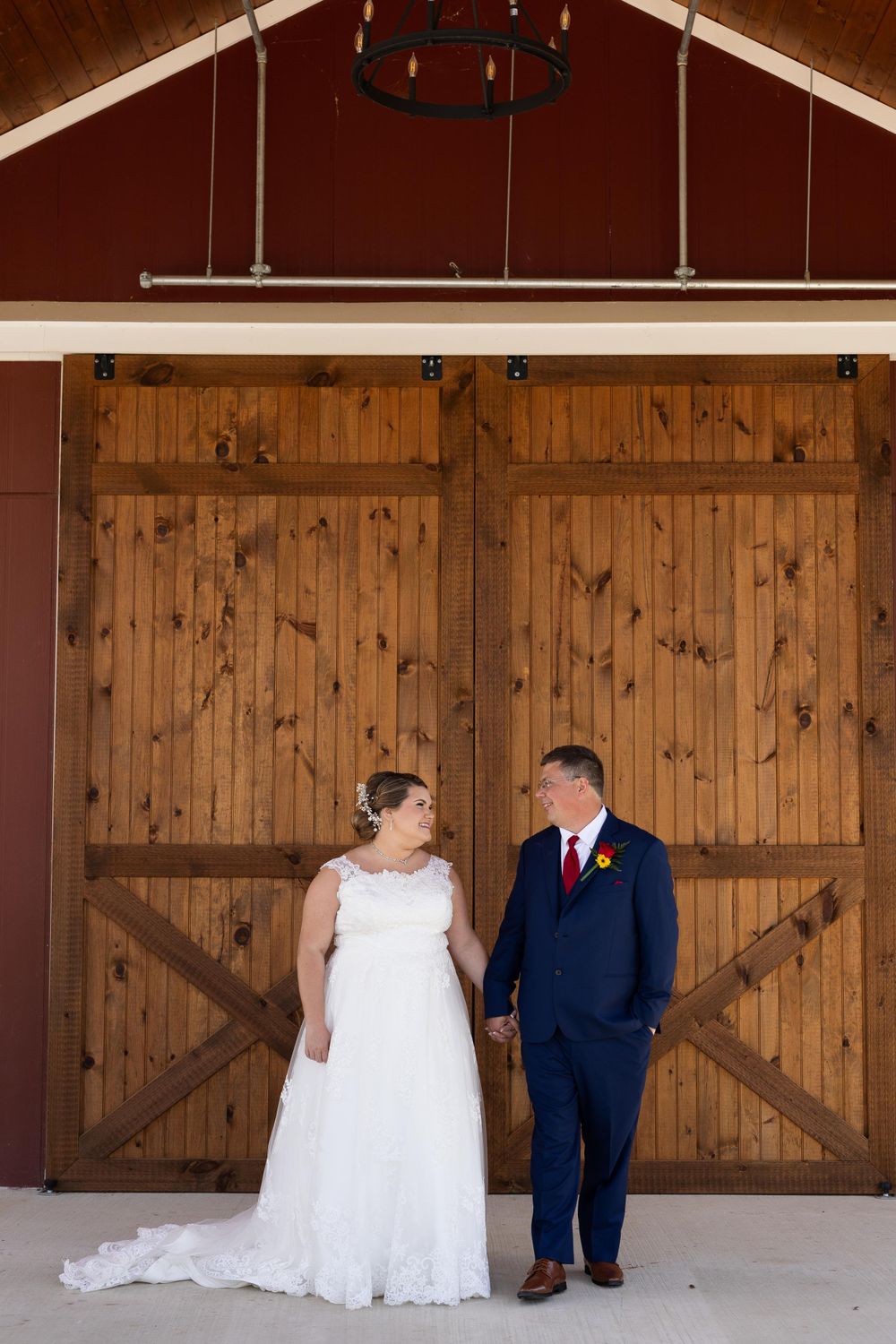 Wedding couple standing against rustic barn doors under a vaulted ceiling with chandelier.