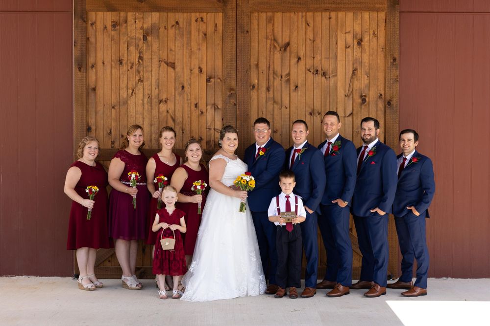 Wedding party poses in burgundy dresses and navy suits against wooden barn door backdrop.